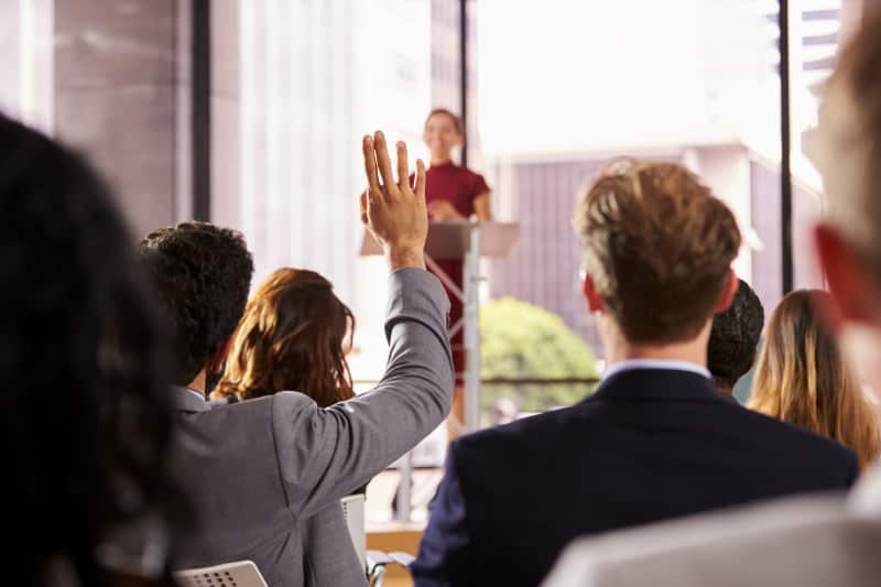 Man with his hand up in audience facing woman at a podium